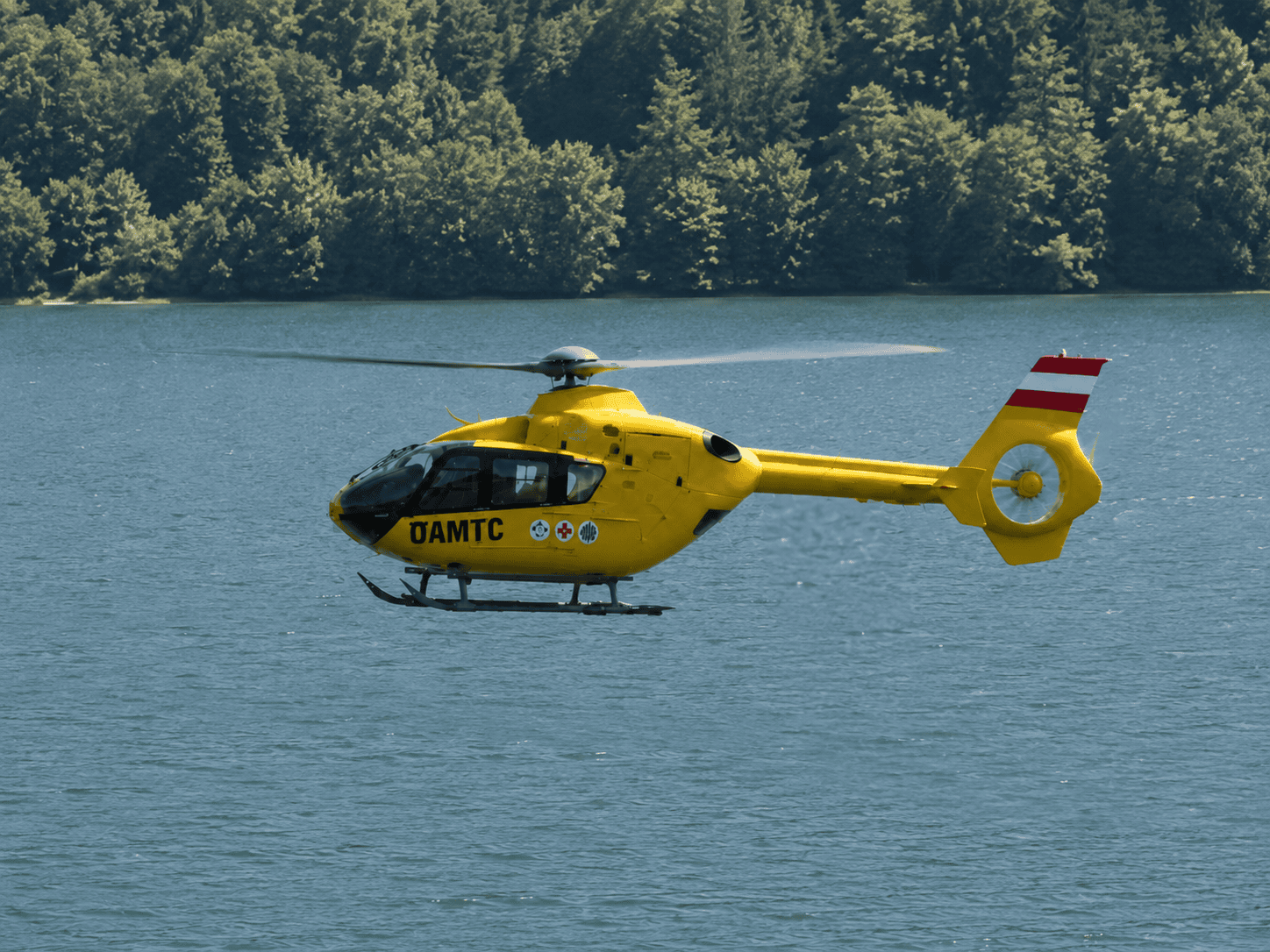 Gelber ÖAMTC-Rettungshubschrauber fliegt über einem See vor dichtem Wald am Ufer