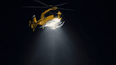 Gelber Rettungshubschrauber mit Suchscheinwerfer im nächtlichen Flug gegen schwarzen Himmel