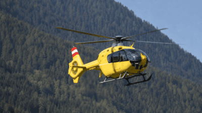 Ein gelber Rettungshubschrauber mit rot-weißem Heck fliegt vor einem dichten Bergwald.