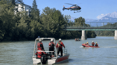 Rettungskräfte in roten Booten auf dem Fluss mit Polizeihubschrauber und Brücke im Hintergrund