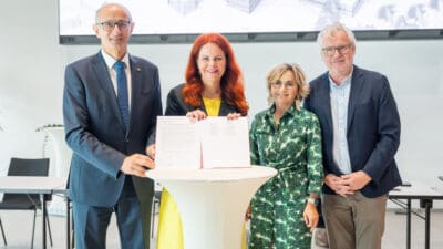 Vier Personen präsentieren ein aufgeschlagenes Dokument auf einem weißen Stehtisch.