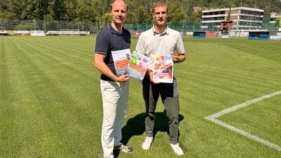 Zwei Männer stehen auf einem grünen Fußballfeld und halten Plakate gegen Gewalt im Sport.