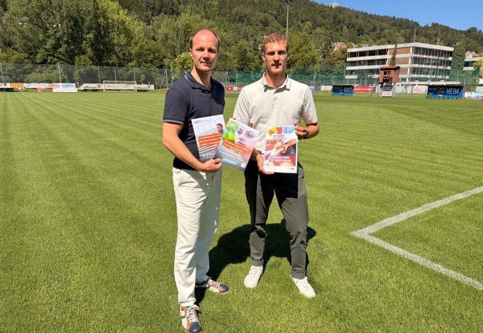 Zwei Männer stehen auf einem grünen Fußballfeld und halten Plakate gegen Gewalt im Sport.
