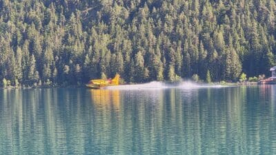 Gelbes Löschflugzeug erzeugt Gischt auf dem türkisfarbenen Weissensee vor bewaldetem Ufer