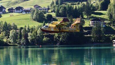 Italienisches Löschflugzeug fliegt über dem Weissensee vor grünen Hügeln und Häusern.