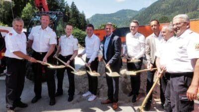 Gruppe von Männern in Uniformen und Anzügen mit Schaufeln beim Spatenstich vor Bagger und Bergen