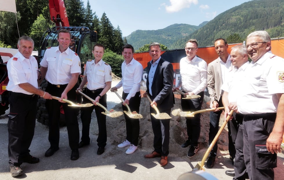 Gruppe von Männern in Uniformen und Anzügen mit Schaufeln beim Spatenstich vor Bagger und Bergen