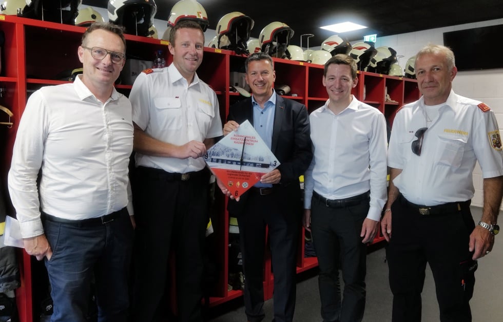 Fünf Männer stehen vor roten Spinden mit Feuerwehrhelmen und halten ein Modell eines Einsatzzentrums.