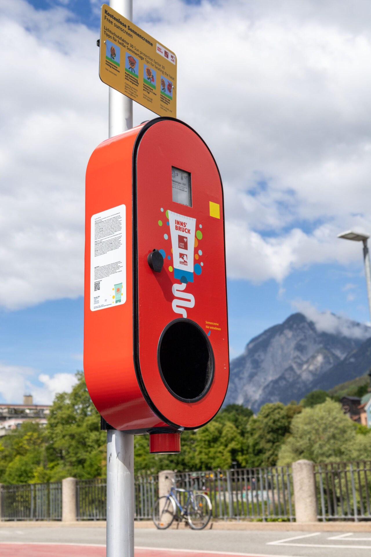 Roter Sonnencremespender mit UV-Anzeige und Hinweisschild an einer Straße in Innsbruck.