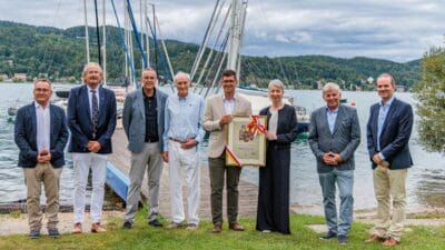 Gruppe von acht Personen hält gerahmtes Kärntner Landeswappen vor Segelbooten am Seeufer