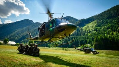 Kniende Soldaten neben einem schwebenden Militärhubschrauber in einer Wiese mit Bergen im Hintergrund.