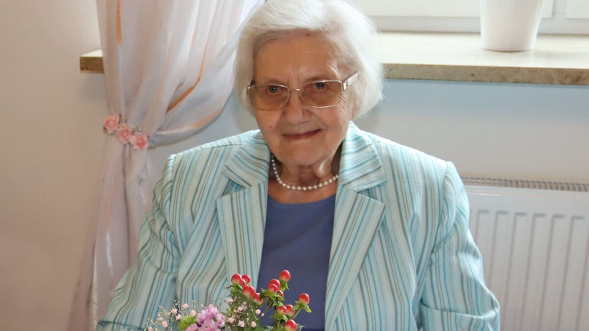 Ältere Frau mit weißem Haar und Brille in gestreiftem Blazer sitzt am Tisch mit Blumenstrauß.