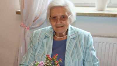 Ältere Frau mit weißem Haar und Brille in gestreiftem Blazer sitzt am Tisch mit Blumenstrauß.