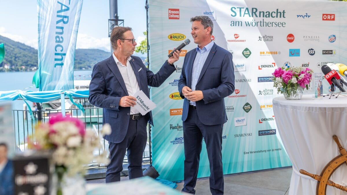 Zwei Männer in Anzügen stehen vor einem großen Banner mit Sponsorenlogos am Wörthersee, einer hält ein Mikrofon.