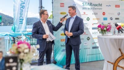 Zwei Männer in Anzügen stehen vor einem großen Banner mit Sponsorenlogos am Wörthersee, einer hält ein Mikrofon.