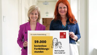 Eine blonde Frau in einem pinken Blazer und eine rothaarige Frau halten gemeinsam ein großes weißes Schild.