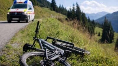 Umgestürztes Mountainbike liegt am grasbewachsenen Straßenrand mit Rettungswagen im Hintergrund in den Bergen