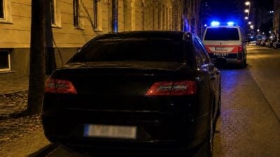 Schwarzes Auto von hinten auf nasser Straße in der Nacht mit Polizeiwagen und Blaulicht im Hintergrund
