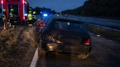 Feuerwehrleute in Warnwesten neben rotem Einsatzfahrzeug und beschädigtem schwarzen Pkw auf nächtlicher Autobahn