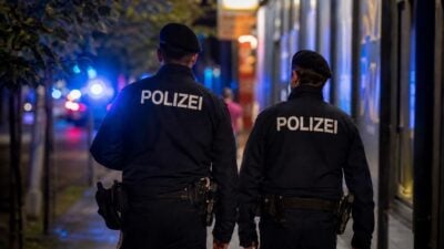 Zwei Polizisten in dunklen Uniformen gehen nachts mit dem Rücken zur Kamera eine Stadtstraße entlang