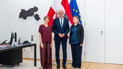 Drei Personen, zwei Frauen und ein Mann, stehen in einem Büro vor mehreren Flaggen und einer Tirol-Karte.