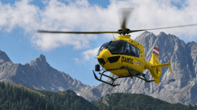 Gelber ÖAMTC-Helikopter fliegt vor einer Berglandschaft mit Wäldern und schroffen Gipfeln.