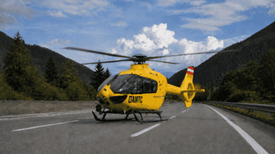 Gelber ÖAMTC-Rettungshubschrauber mit laufenden Rotoren auf einer Autobahn vor Bergen