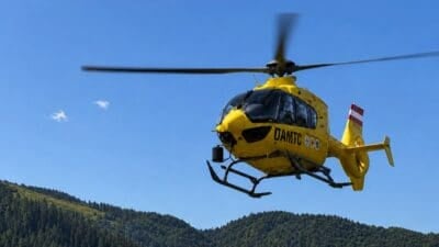 Gelber ÖAMTC-Rettungshubschrauber fliegt über bewaldete Berge unter blauem Himmel.