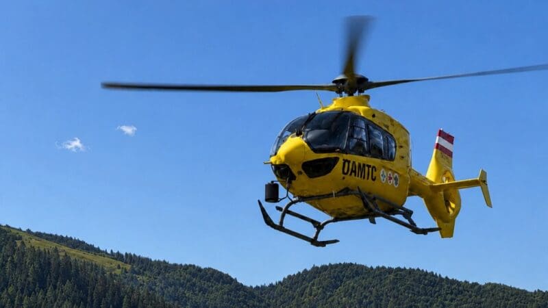 Gelber ÖAMTC-Rettungshubschrauber fliegt über bewaldete Berge unter blauem Himmel.
