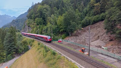 Roter Zug fährt an Baustelle mit Baggern durch bewaldete Berglandschaft.