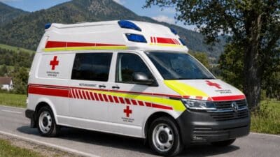 Weißer Rettungswagen mit rotem Kreuz und blauen Lichtern auf einer Straße vor den Alpen
