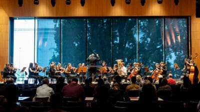 Dirigent dirigiert Orchester mit Streichern und Blechbläsern vor Publikum in hellem Konzertsaal mit Fenstern