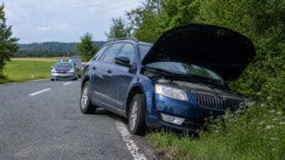 Dunkelblaues Auto mit offener Motorhaube am Straßenrand und Polizeifahrzeug mit Blaulicht im Hintergrund