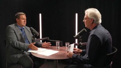 Zwei Männer in Anzügen sitzen an einem Tisch mit Mikrofonen und Papieren in einem dunklen Studio.