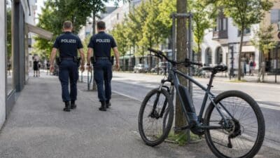 Zwei Polizisten in Uniform gehen auf einem Gehweg an einem E-Bike vorbei, das an einem Baum befestigt ist.