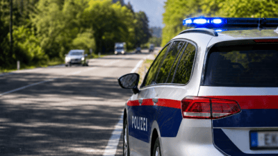 Heckansicht eines silbernen Polizeiautos mit rot-blauem Streifen und blauem Blaulicht auf einer Landstraße.