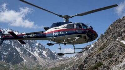Polizeihubschrauber mit Kennung OE-BXL fliegt vor schneebedeckten Alpenbergen unter blauem Himmel