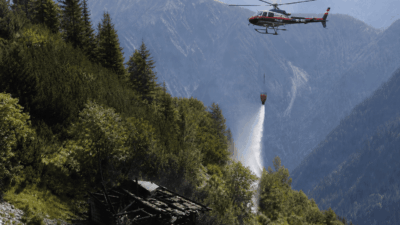 Polizeihubschrauber lässt Wasser aus einem Behälter auf ein Feuer in den Bergen fallen