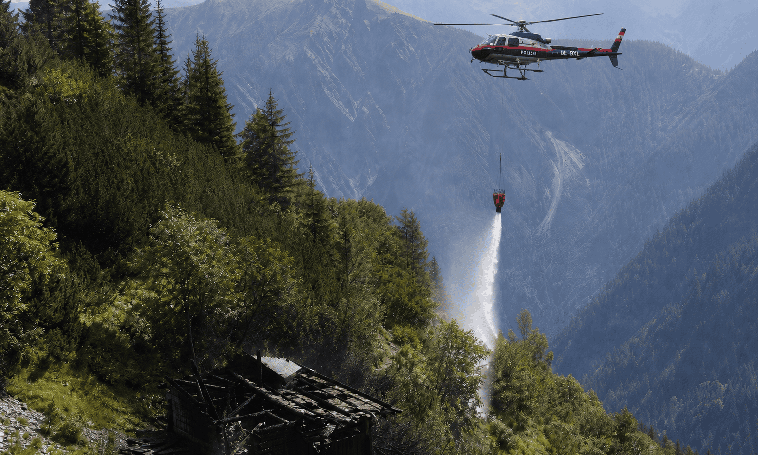 Polizeihubschrauber lässt Wasser aus einem Behälter auf ein Feuer in den Bergen fallen