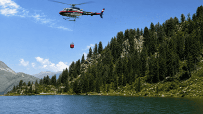 Polizeihubschrauber mit rotem Löschbehälter fliegt über einem alpinen Bergsee in bewaldeter Berglandschaft