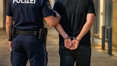 Ein Polizist in blauer Uniform führt einen Mann mit Handschellen hinter dem Rücken auf dem Gehweg.