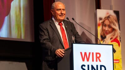 Älterer Mann in dunklem Anzug und roter Krawatte spricht an einem Podium mit der Aufschrift WIR SIND.