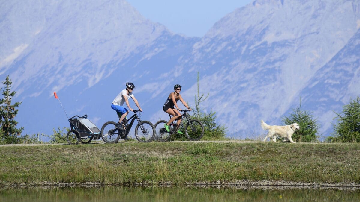 Mann und Frau auf Fahrrädern mit Hund an einem See vor Bergen in Tirol