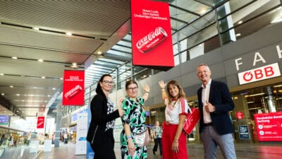 Vier Personen posieren vor einer großen roten Coca-Cola-Werbetafel im Wiener Hauptbahnhof.