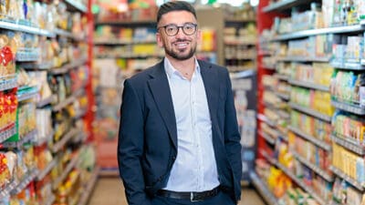 Lächelnder Mann mit Brille und Bart im Anzug zwischen Supermarktregalen