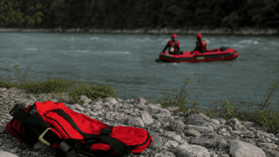 Roter Rettungsrucksack auf steinigem Ufer vor einem Schlauchboot mit zwei Helfern auf dem Fluss