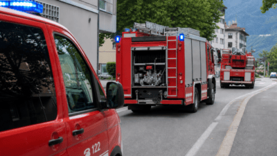 Rote Feuerwehrfahrzeuge mit Blaulicht auf einer Straße vor Gebäuden und Bergen