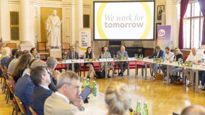 Teilnehmer sitzen an einem U-förmigen Tisch in einem prächtigen Saal mit Statue und großer Leinwand.