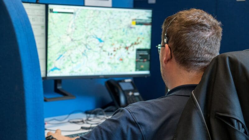 Rückansicht eines Mannes mit Headset vor einem Monitor mit geografischer Karte in einem Kontrollraum