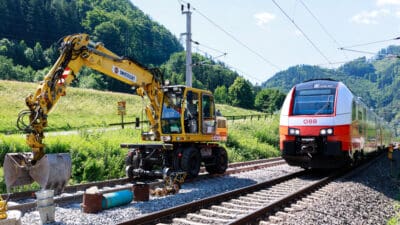 Gelber Schienenbagger von Swietelsky neben rot-weißem ÖBB-Zug auf Bahngleisen